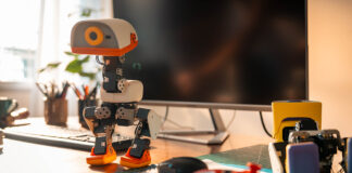MicroDuck biped robot standing on a desk beside a computer and game controller