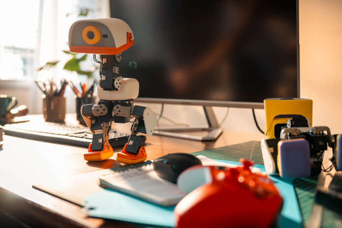 MicroDuck biped robot standing on a desk beside a computer and game controller