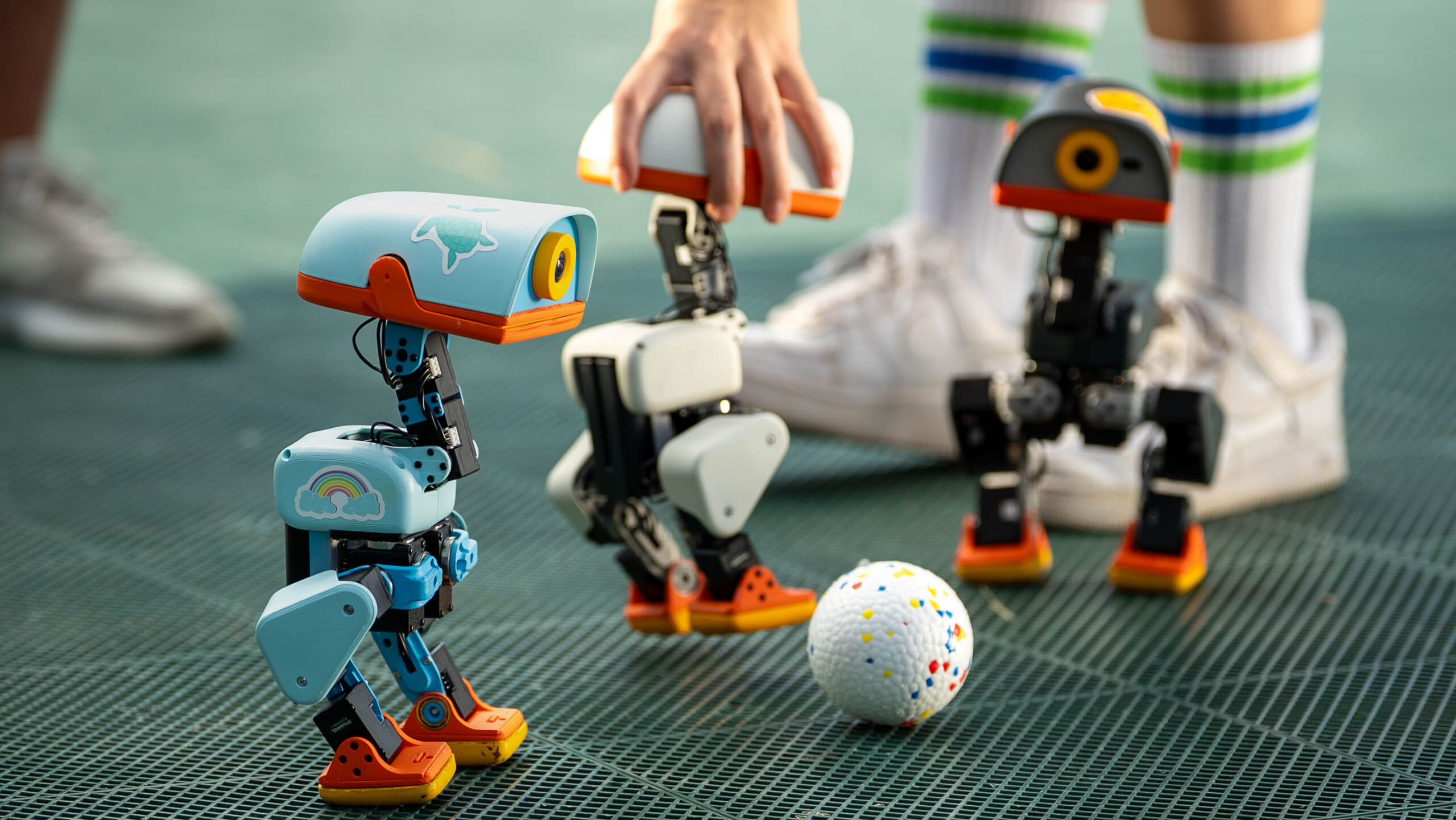 Three colorful MicroDuck biped robots standing around a small ball