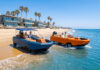 Blue and orange WaterCar EV amphibious vehicles driving out of shallow ocean water onto a beach